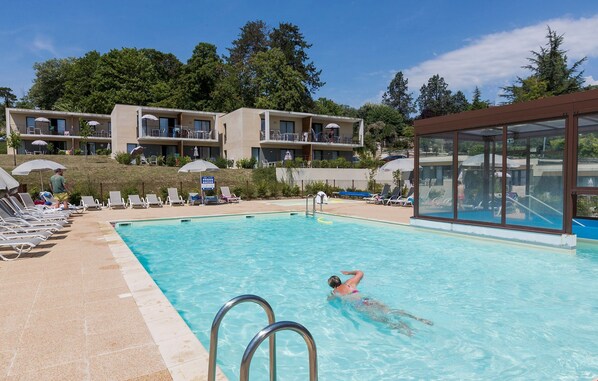 Una piscina con sillones de descanso y sombrillas, un espacio cubierto de vidrio y un edificio con balcones al fondo.