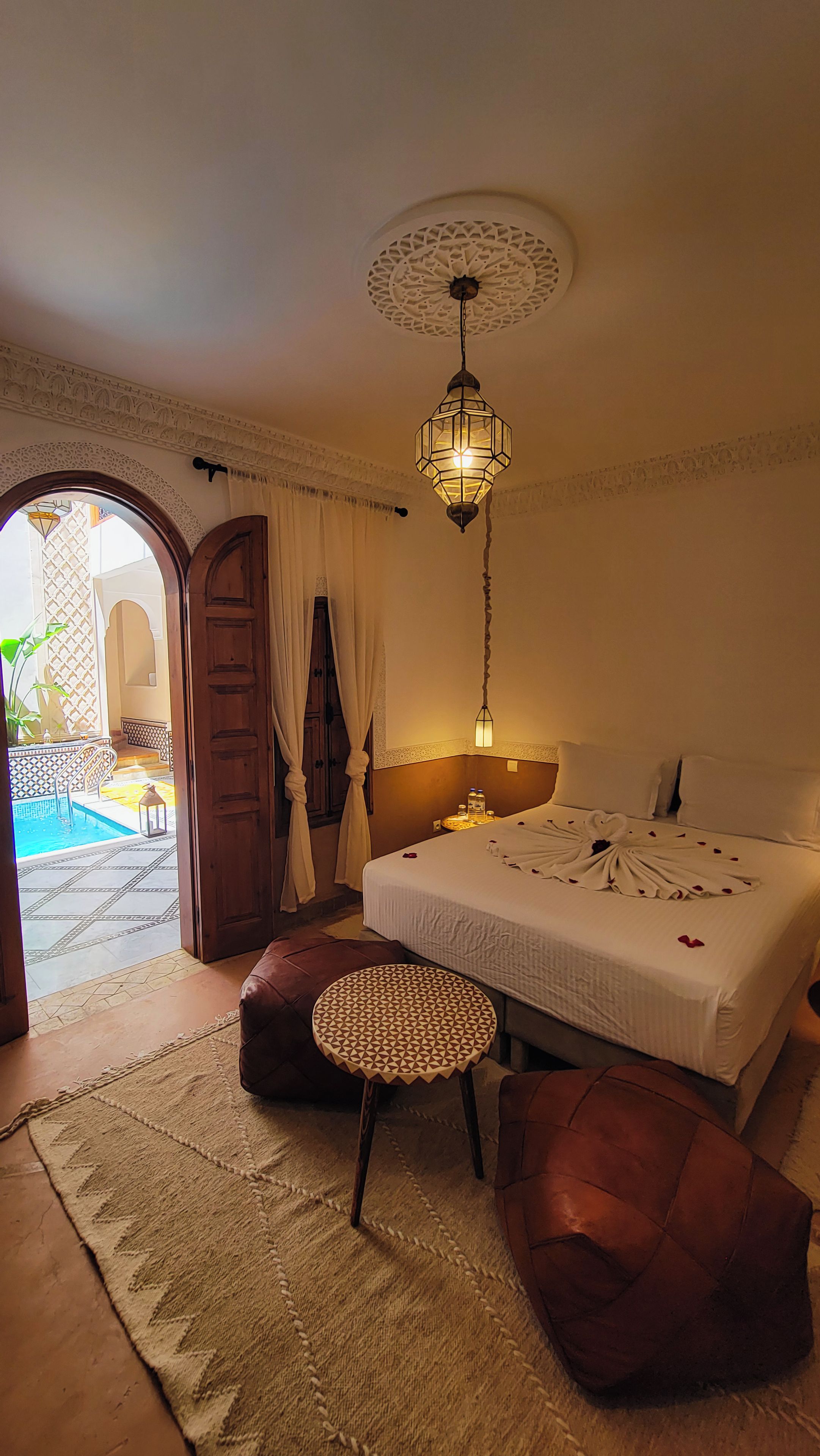 A hotel room with a bed, a round table, a hanging light fixture, and a view of an outdoor swimming pool.