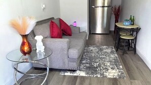 A modern living room with a grey sofa, a glass coffee table, a stainless steel refrigerator, and a small round table with chairs.