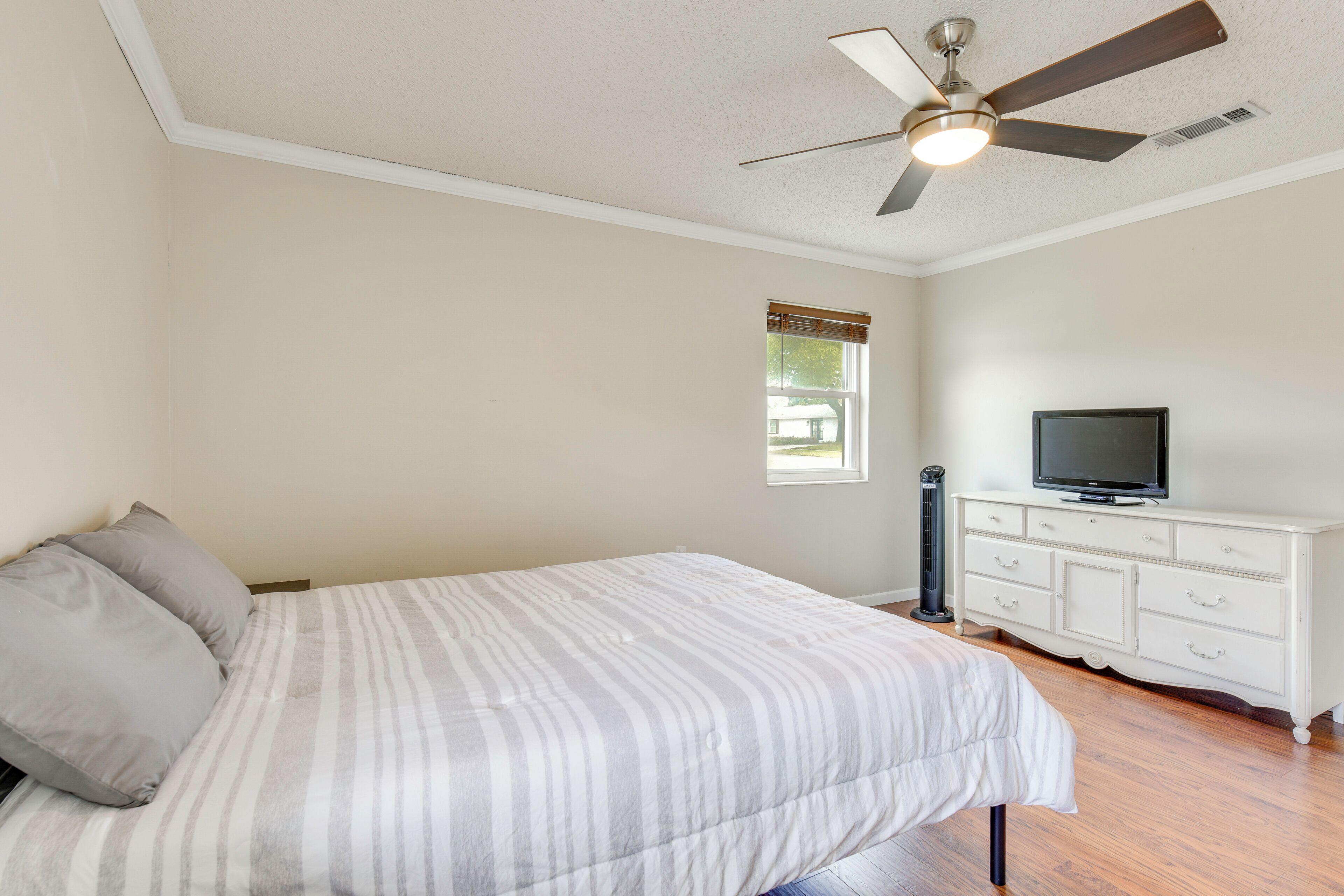 Une chambre à coucher avec un lit, un téléviseur posé sur une commode, un ventilateur de plafond et une fenêtre.