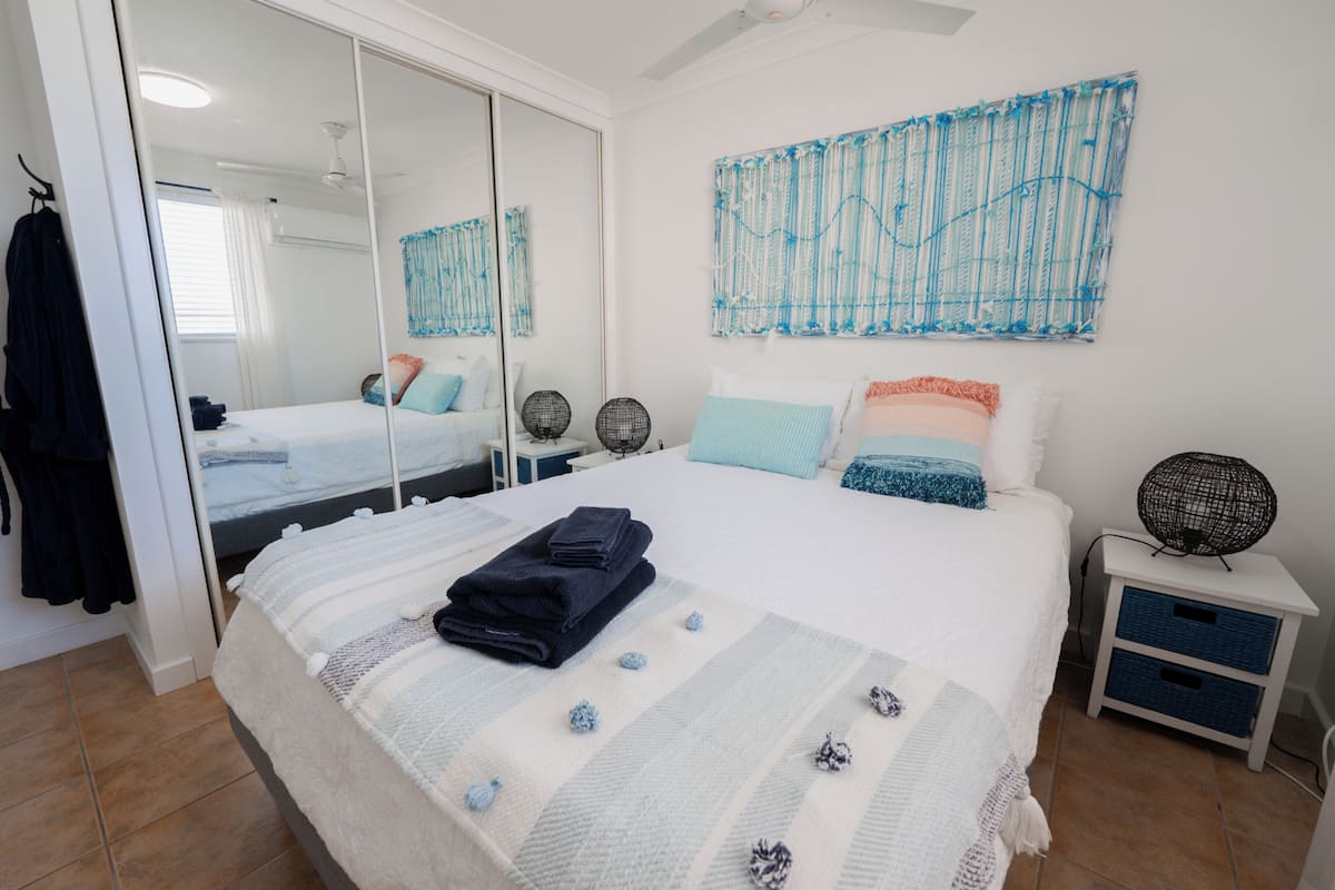 A bedroom with a bed, a bedside table with a fan, a wall-mounted artwork, and a large mirror.