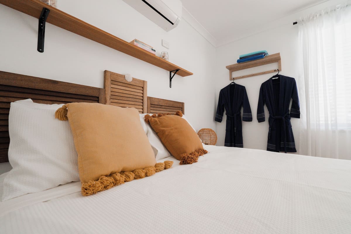 A neatly made bed with two pillows, wooden headboards, and a wall-mounted shelf with items.