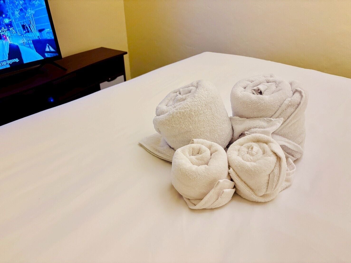 Una cama bien hecha, toallas enrolladas y un televisor al fondo.