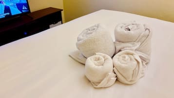 Una cama bien hecha, toallas enrolladas y un televisor al fondo.