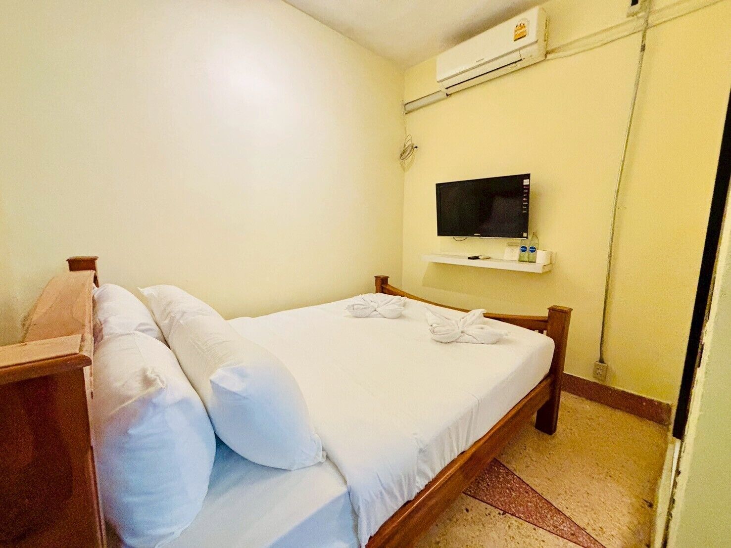 Una cama individual con ropa de cama y almohadas blancas, un televisor montado y un equipo de aire acondicionado en la pared.