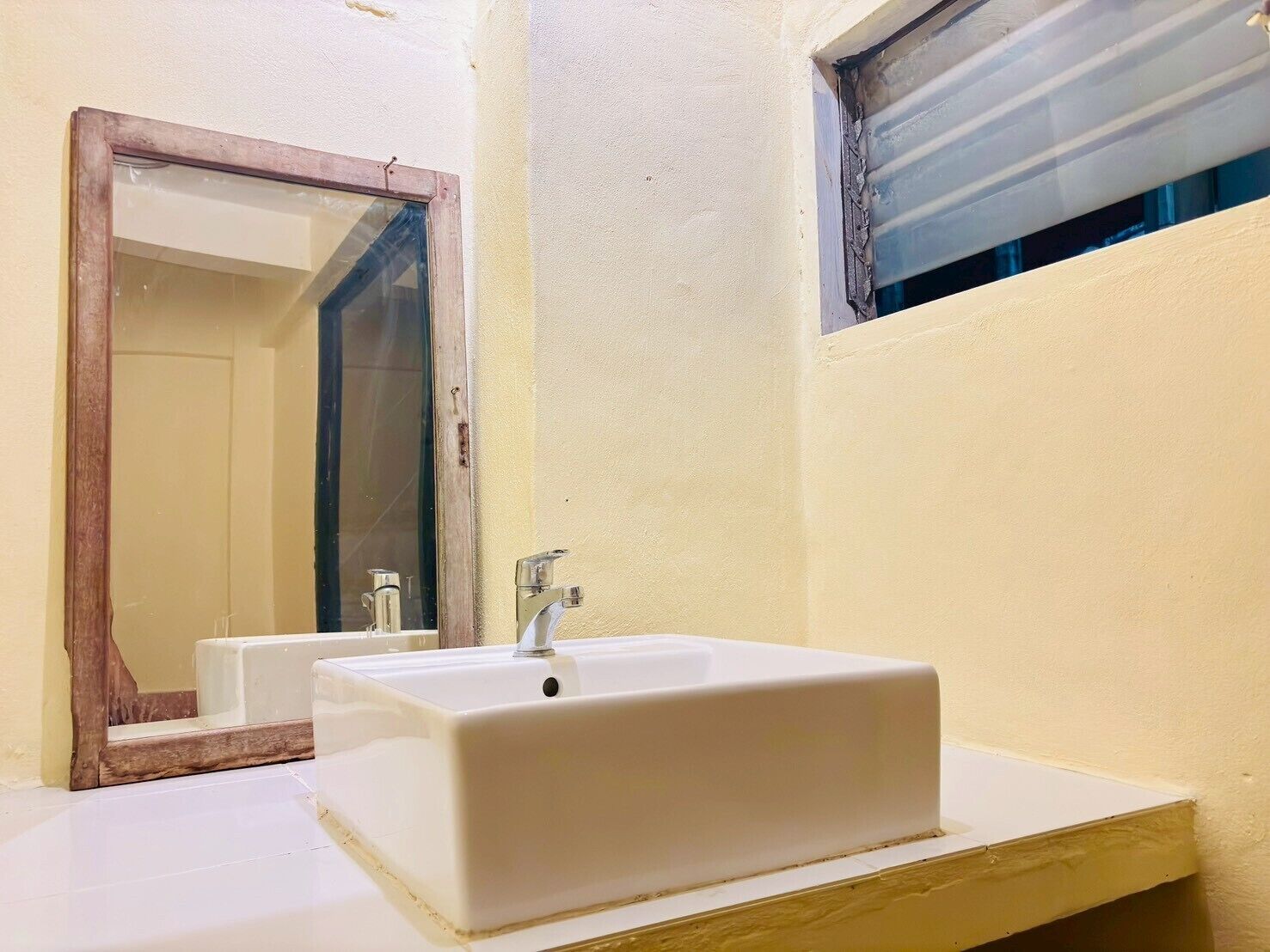 Un baño con lavamanos blanco, un espejo y una ventana con persianas.