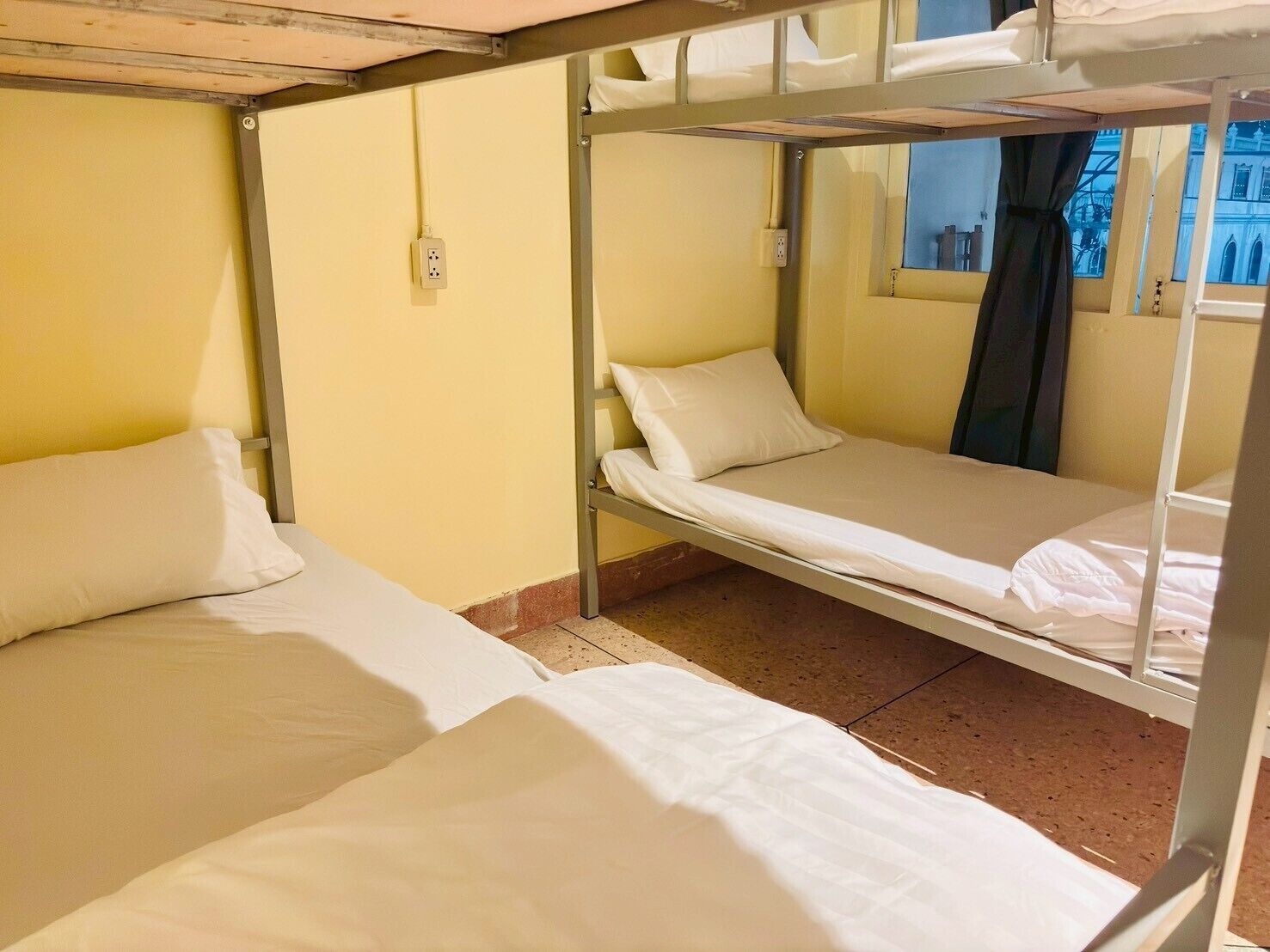 A dormitory room with bunk beds, a window with a curtain, and a visible electrical outlet.