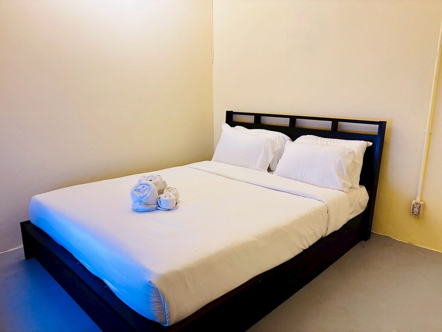 A neatly made bed with white linens and pillows, a rolled towel, and a power outlet on the wall.