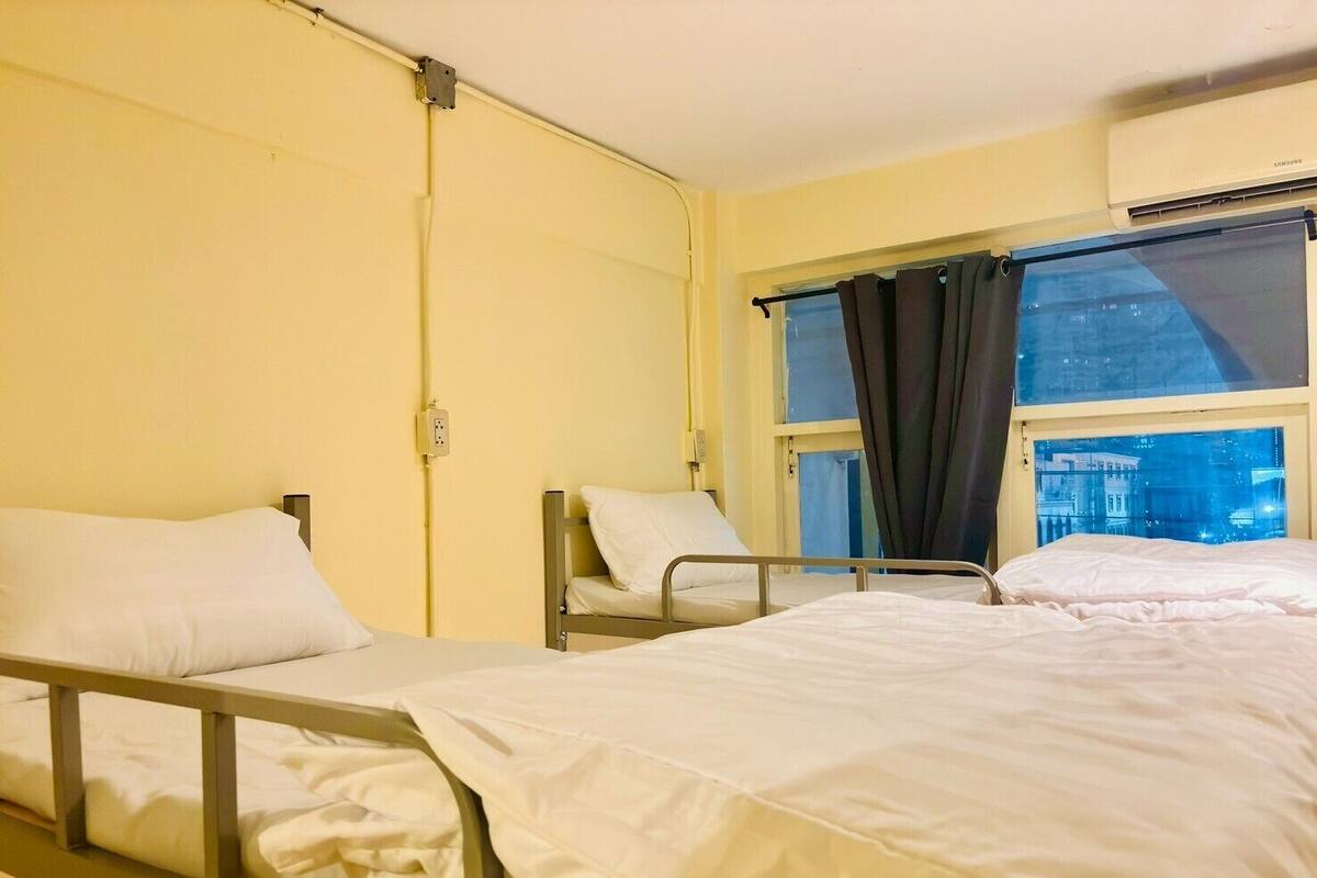 A hospital room with two beds, a window with curtains, an air conditioning unit, and a wall-mounted electrical outlet.