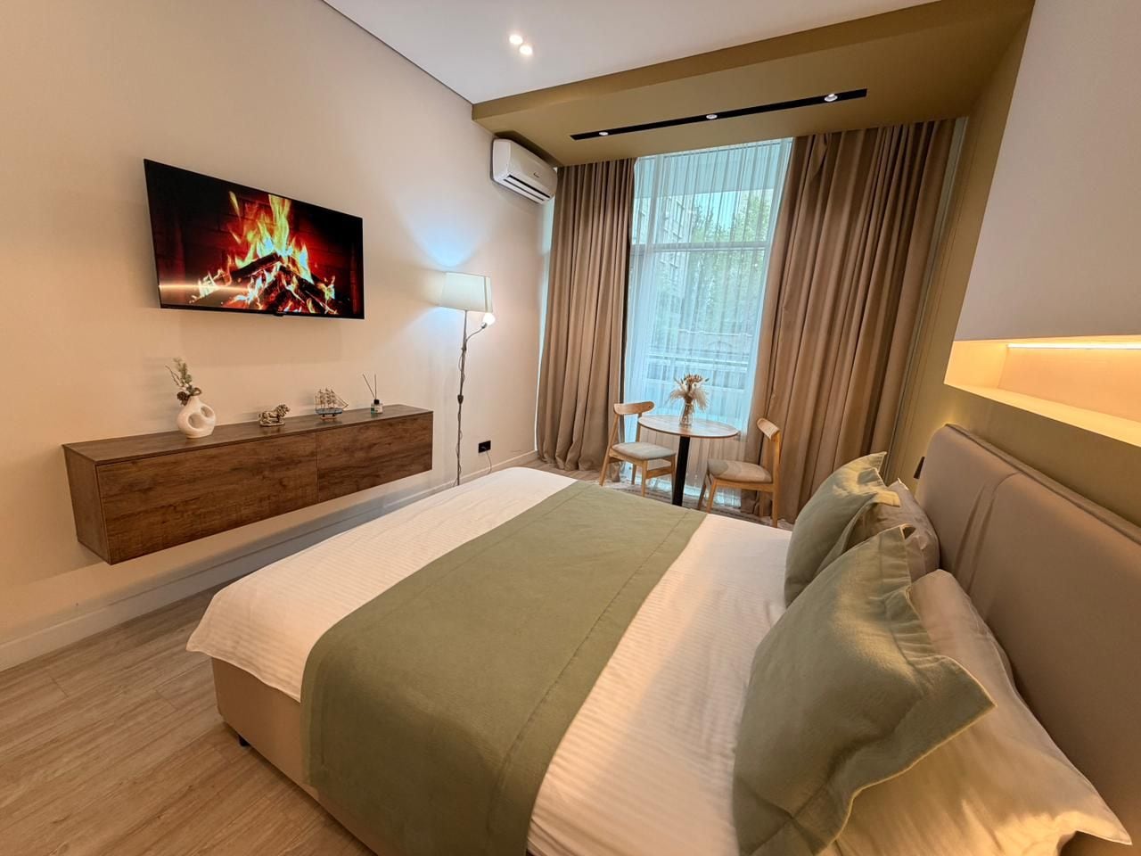 Une chambre d’hôtel moderne équipée d’un lit, d’un téléviseur fixé au mur, d’une table de chevet, d’une lampe et d’une fenêtre avec des rideaux.