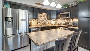 Una cocina moderna con electrodomésticos de acero inoxidable, armarios oscuros, encimera de granito e iluminación colgante.