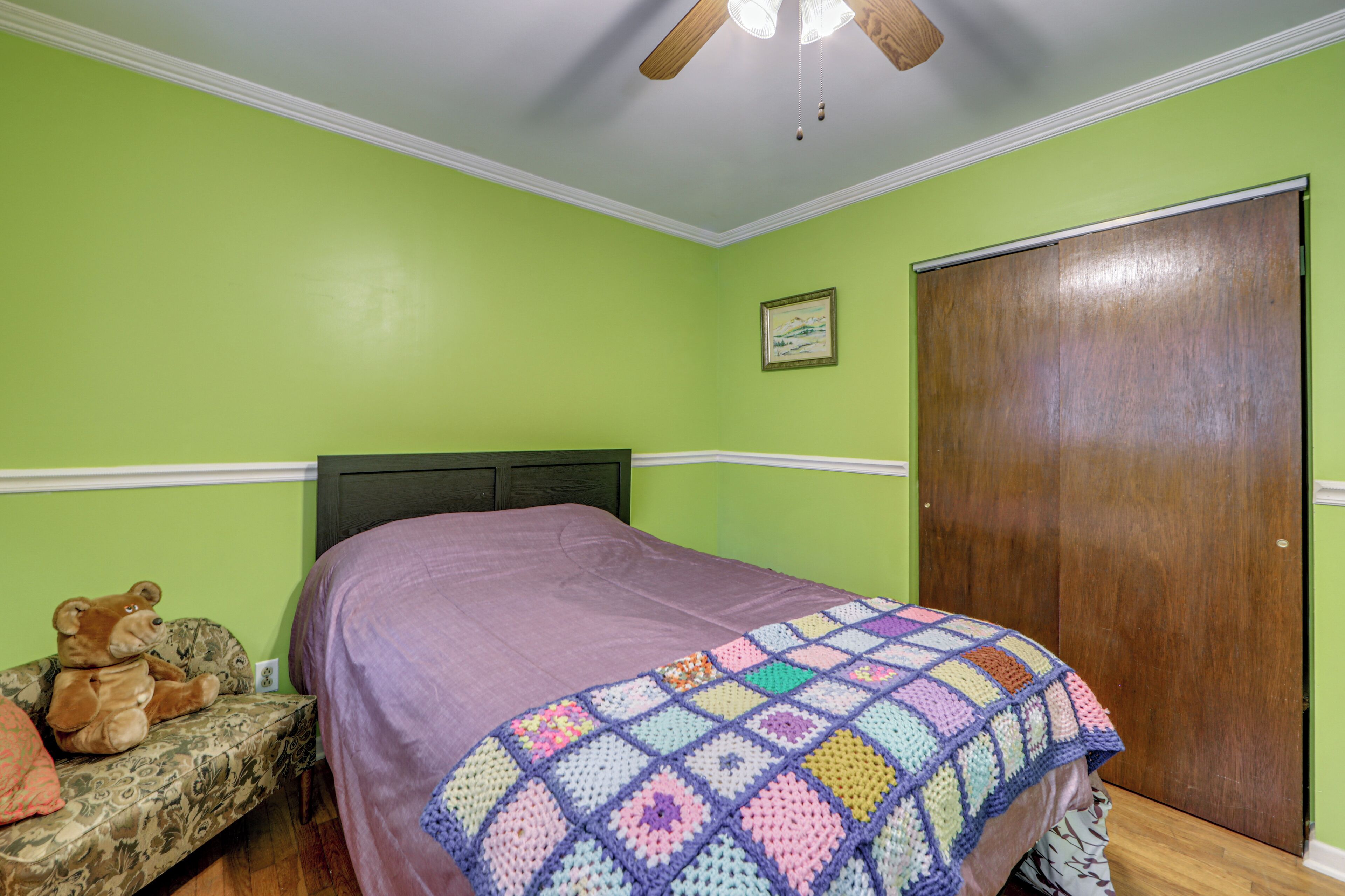 Une chambre à coucher avec un lit, un ours en peluche, une courtepointe, une porte en bois et un ventilateur de plafond.