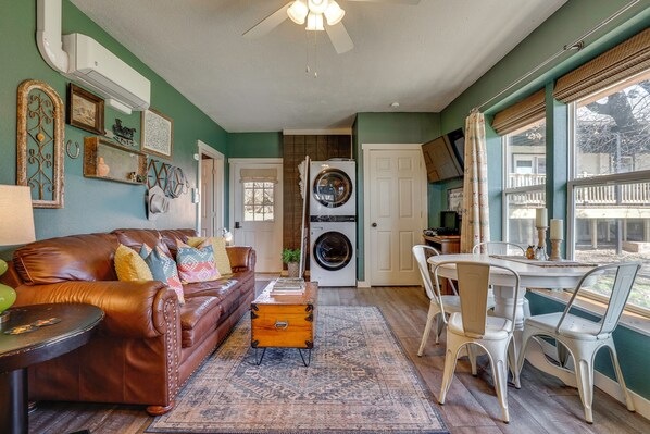 A living room with a brown leather couch, a wooden chest, and a dining area with white chairs.