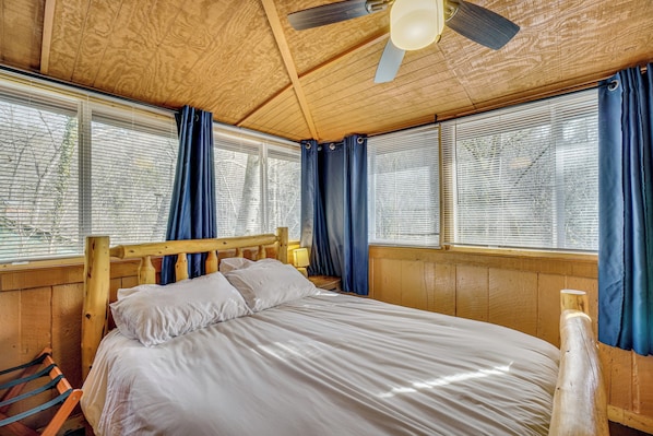 Una habitación con estructura de madera, una cama, ventanas con cortinas y un ventilador de techo.