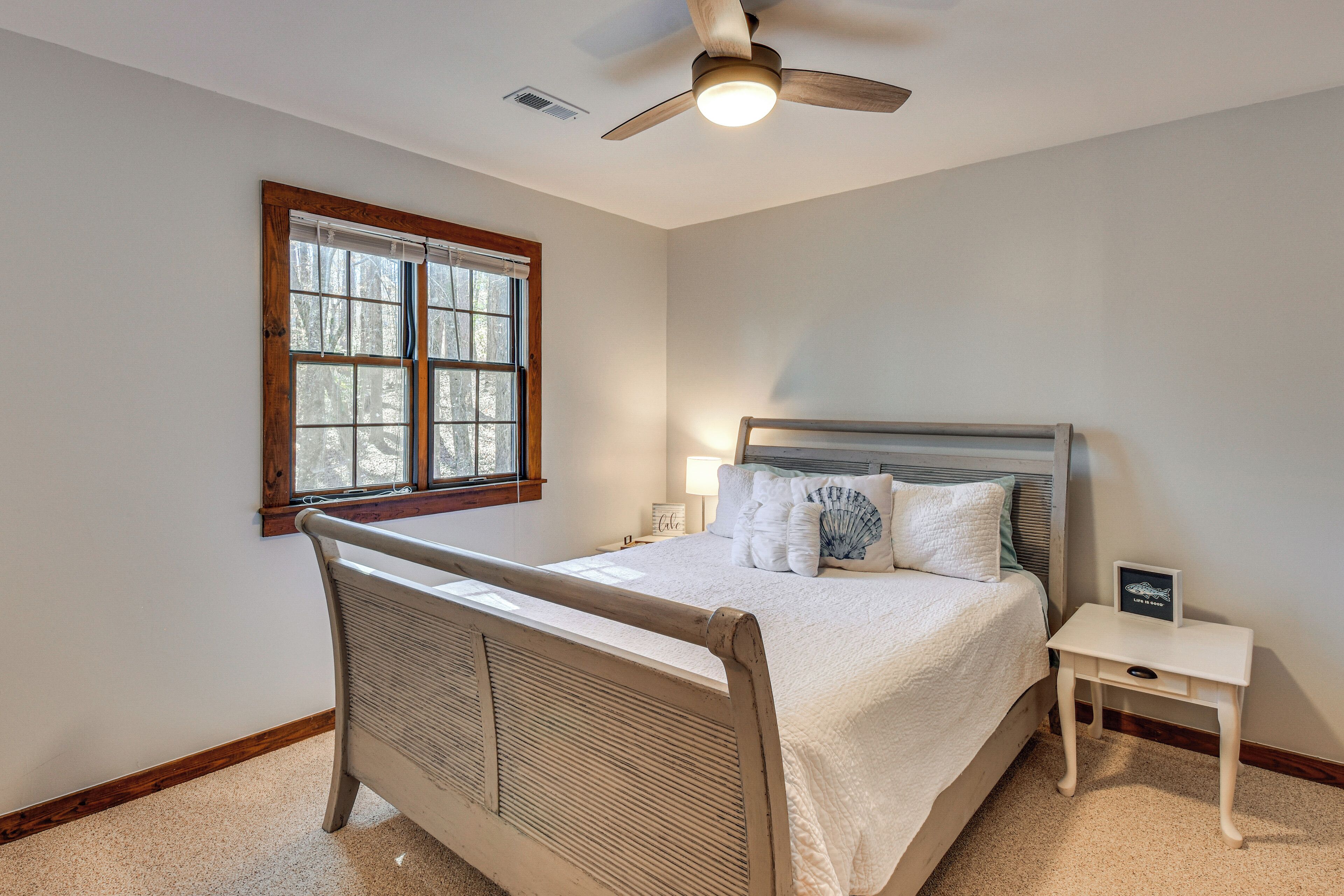 A bedroom with a bed, a nightstand, a ceiling fan, a window, and a wall-mounted light fixture.