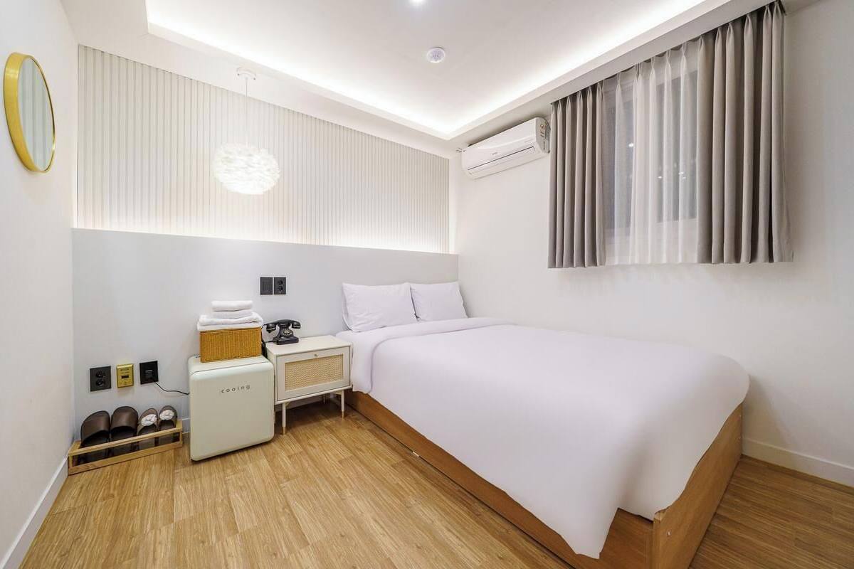 Une chambre d’hôtel moderne équipée d’un lit, d’une table de chevet, d’une lampe, d’un climatiseur et d’un petit coin salon avec un panier et un téléphone.