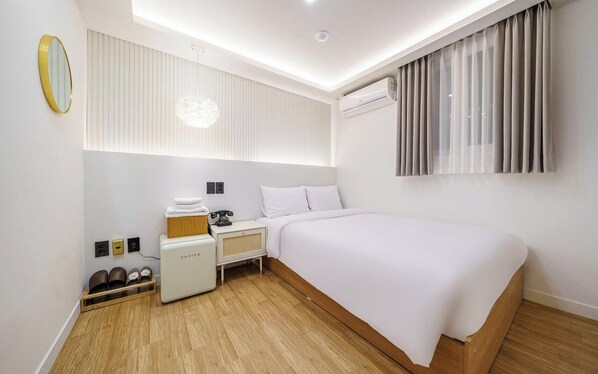 Une chambre d’hôtel moderne équipée d’un lit, d’une table de chevet, d’une lampe, d’un climatiseur et d’un petit coin salon avec un panier et un téléphone.