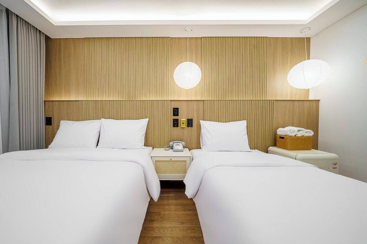 Une chambre d’hôtel avec deux lits, une tête de lit en bois, des tables de chevet avec des lampes et un petit panier posé sur la table de chevet.