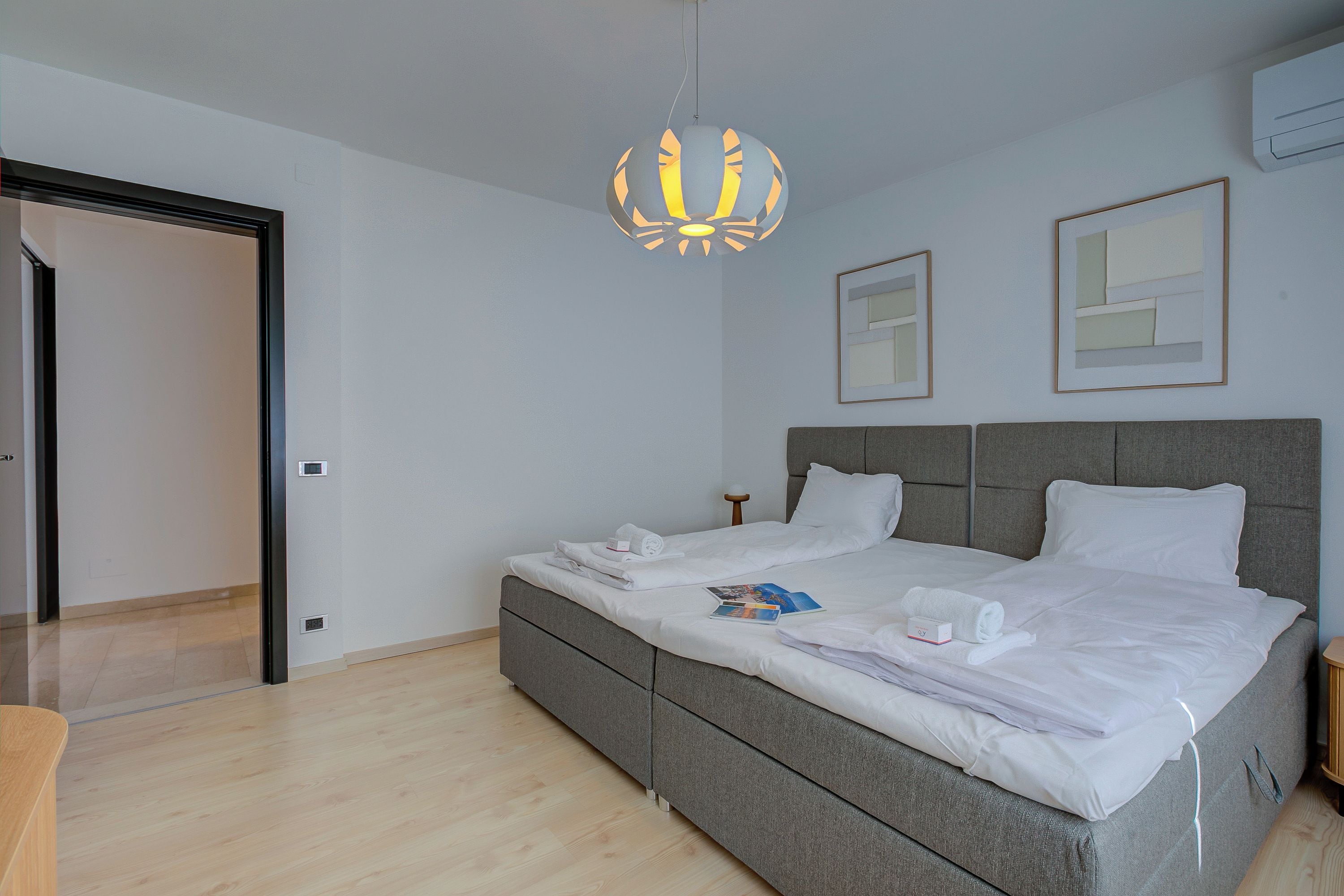 A modern bedroom with a grey bed, white linens, a unique pendant light, and two framed pictures on the wall.