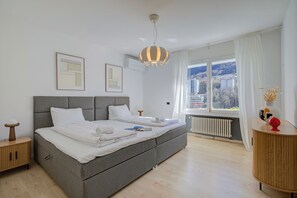 A modern bedroom with a large bed, a bedside table, a dresser, and a view of the city through the window. - Lugano Lake View Villa w. Private Pool (Lugano)