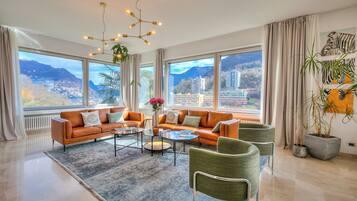 A modern living room with a sofa set, coffee table, and chairs, large windows with a mountain view, and a hanging light fixture.