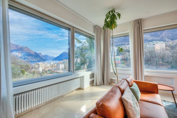 A modern living room with a leather sofa, large windows, and a view of mountains and buildings. - Lugano Lake View Villa w. Private Pool (Lugano)