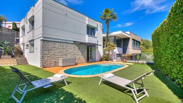 A modern two-story house with a swimming pool, stone facade, and a well-maintained lawn.