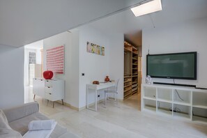 A modern living room with a white desk, a clear chair, a sofa, a TV, and a bookshelf.