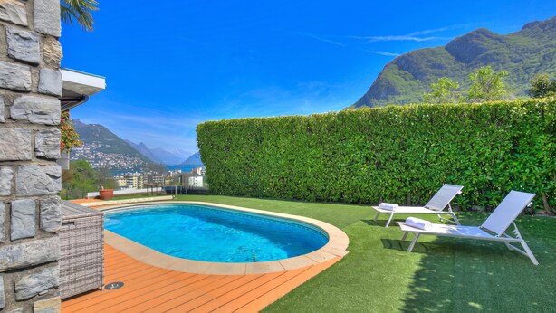 Una piscina con cubierta de madera, dos sillas reclinables y un seto.