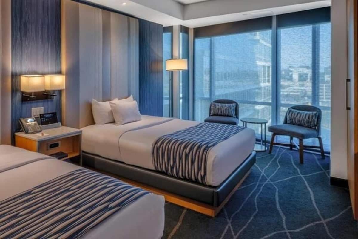 Habitación de hotel con dos camas, una mesita, sillas y un ventanal con vistas.