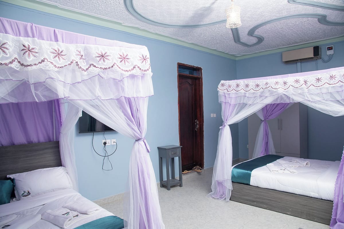 A room with two beds, each with a canopy, a small table, a wall-mounted TV, and a door.