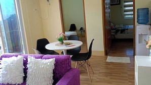Een kamer met een paarse bank, een eettafel met stoelen, een waterdispenser en een grote spiegel.
