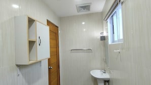 Een badkamer met een houten deur, een wit kastje, een wastafel en een raam met luxaflex.