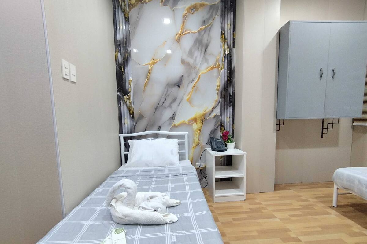 Een opgemaakt bed met een handdoek, een klein nachtkastje met een telefoon en een potplant, een kast en een marmeren muur.