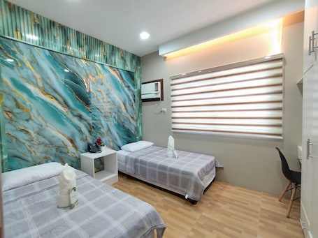 A hotel room with two beds, a small bedside table with a phone and a flower, a wall with a marble-like design, a window with blinds, and a chair.