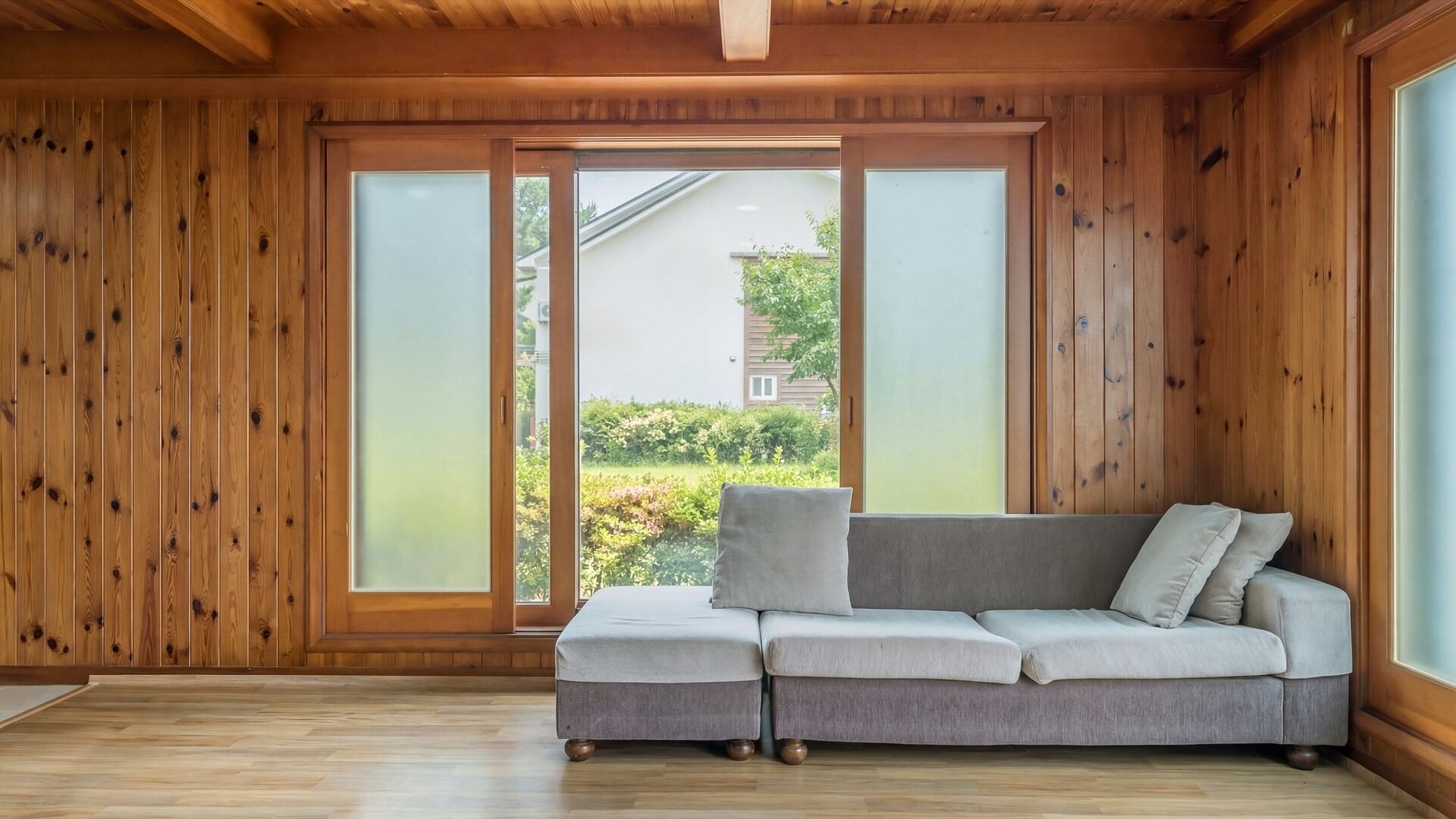 A room with wooden walls, a grey sofa, and a sliding glass door leading to a garden.