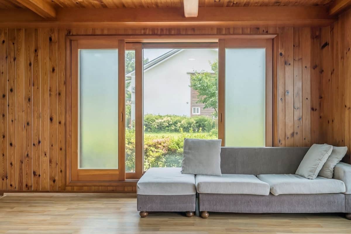 Ein Zimmer mit Holzwänden, einem grauen Sofa und einer Schiebetür zu einem Garten.