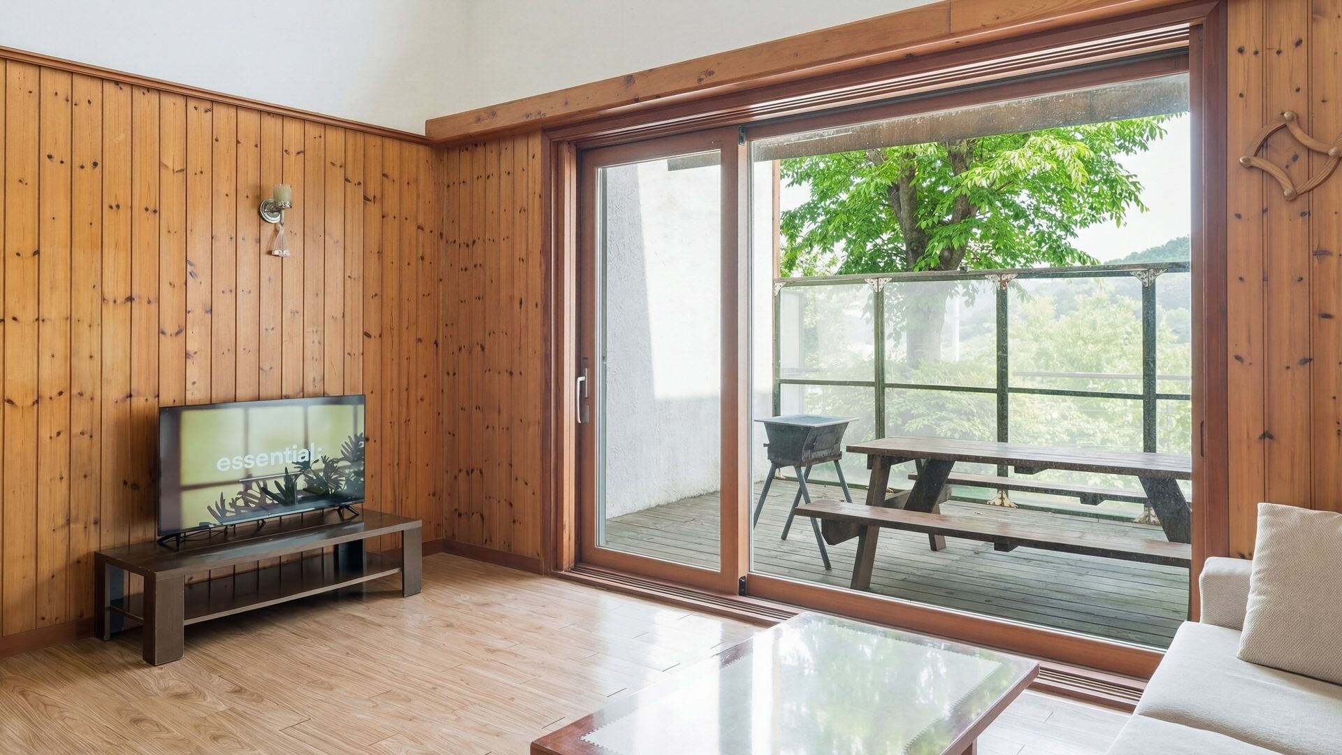 A room with wooden walls, a sliding glass door leading to an outdoor deck, a television on a stand, and a glass coffee table.