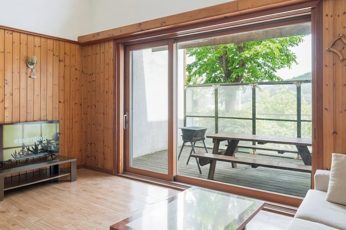 Ein Zimmer mit Holzwänden, einer Schiebetür zu einem Terrassenbereich, einem Fernseher auf einem Beistelltisch und einem Glastisch.