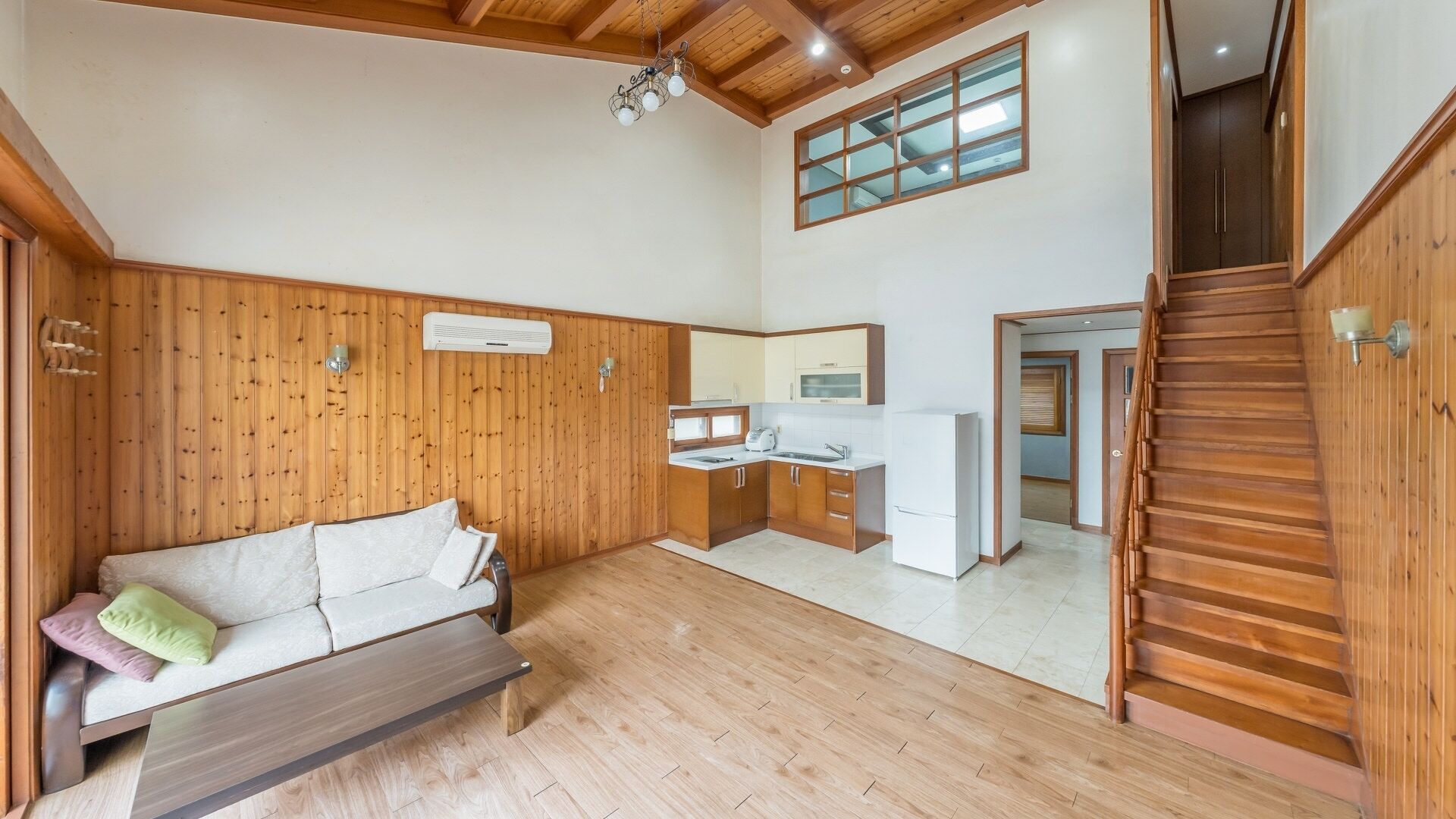 A spacious room with a wooden floor, a sofa, a kitchen area with a refrigerator, and a staircase.