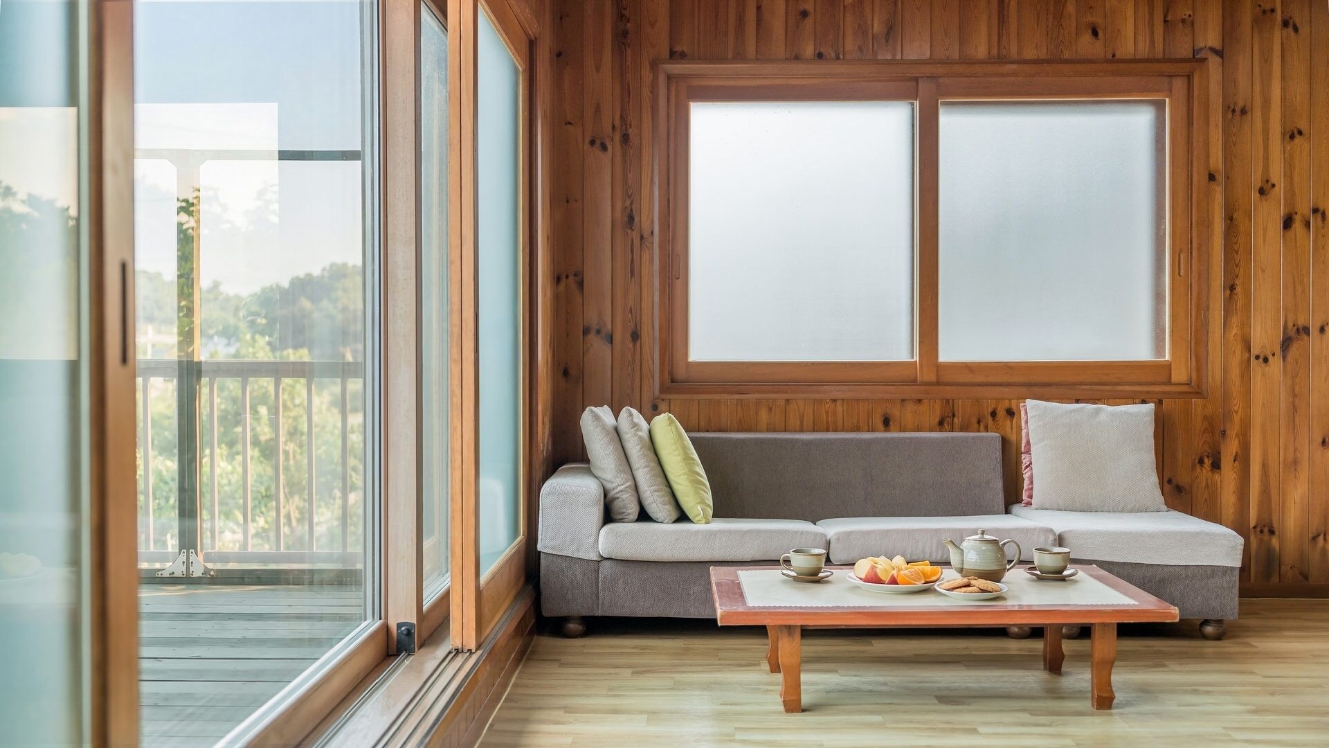 A room with a wooden paneled wall, a gray sofa with cushions, a wooden coffee table with a teapot and cups, and a view through large windows.