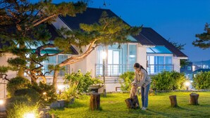 Ein modernes Haus mit einem großen Glasfenster, einem gepflegten Garten und einer Person, die einen Baum pflegt.