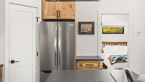 Una cucina compatta con piano cottura a gas, frigorifero in acciaio inox, mobili in legno e un piccolo cassetto in legno, oltre a un letto con vista su una finestra.