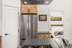 A compact kitchen with a gas stove, stainless steel refrigerator, wooden cabinets, a small wooden drawer, and a bed with a window view. - 2BR New Home in Twin Lakes, Views, Pet Friendly (Twin Lakes)