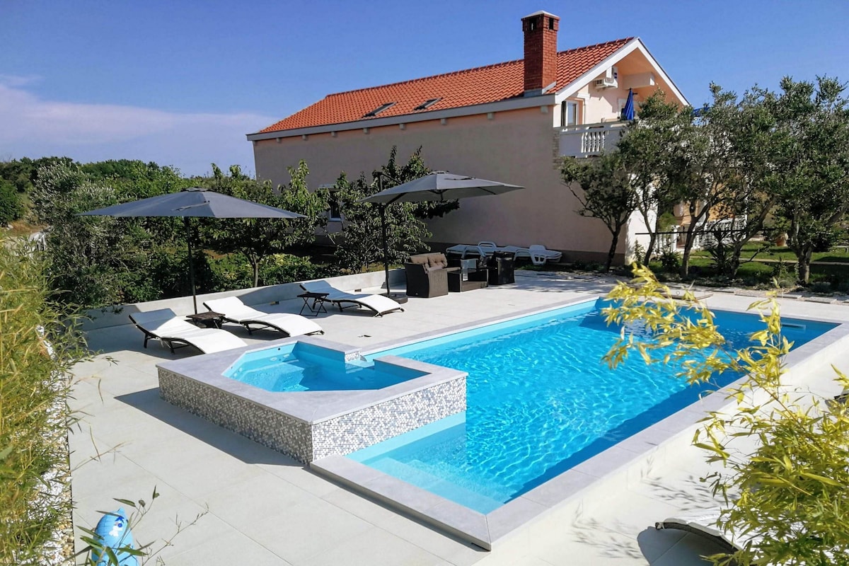 A swimming pool with lounge chairs and umbrellas, a hot tub, and a house in the background.