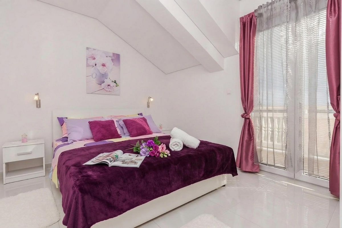 A bedroom with a bed, pillows, a nightstand, a window with curtains, and a wall-mounted light.