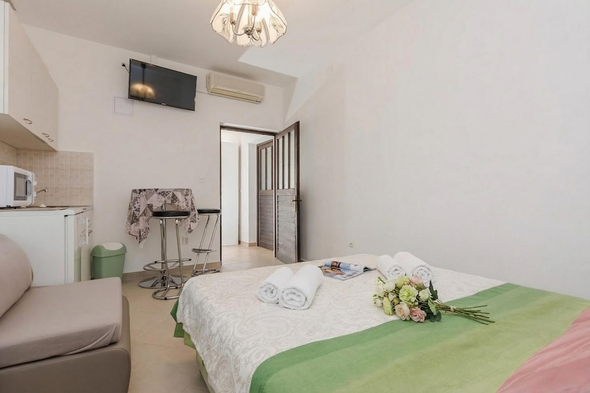 A hotel room with a bed, a TV, a small kitchenette, and a bathroom visible through an open door.