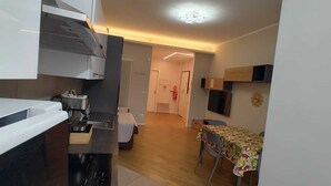 Eine kleine Küche mit modernen Geräten, ein Essbereich mit Tisch und Stühlen sowie ein Flur, der zum Schlafzimmer führt.