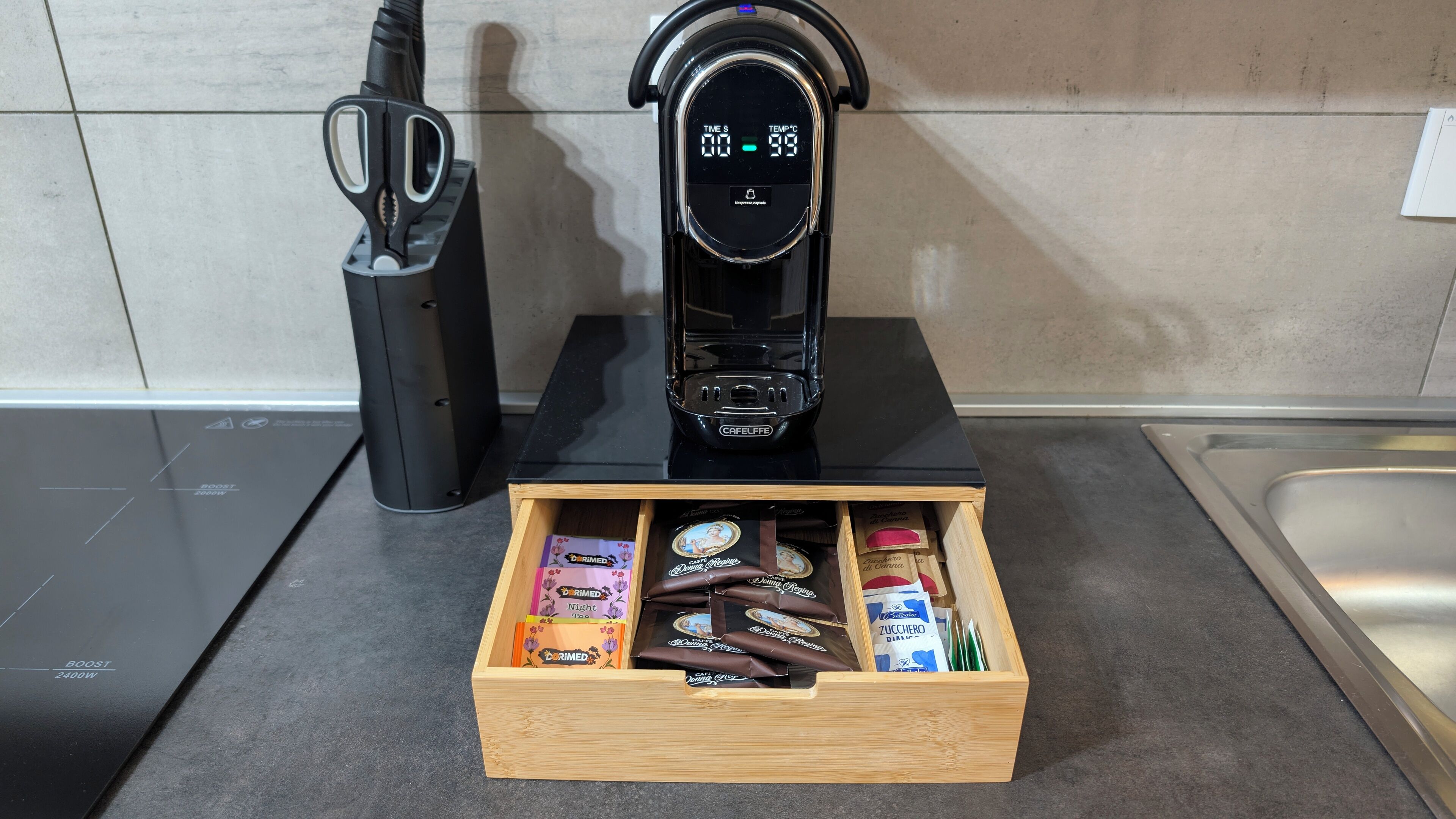 A kitchen with a coffee maker, a drawer of coffee pods, and a pair of scissors.