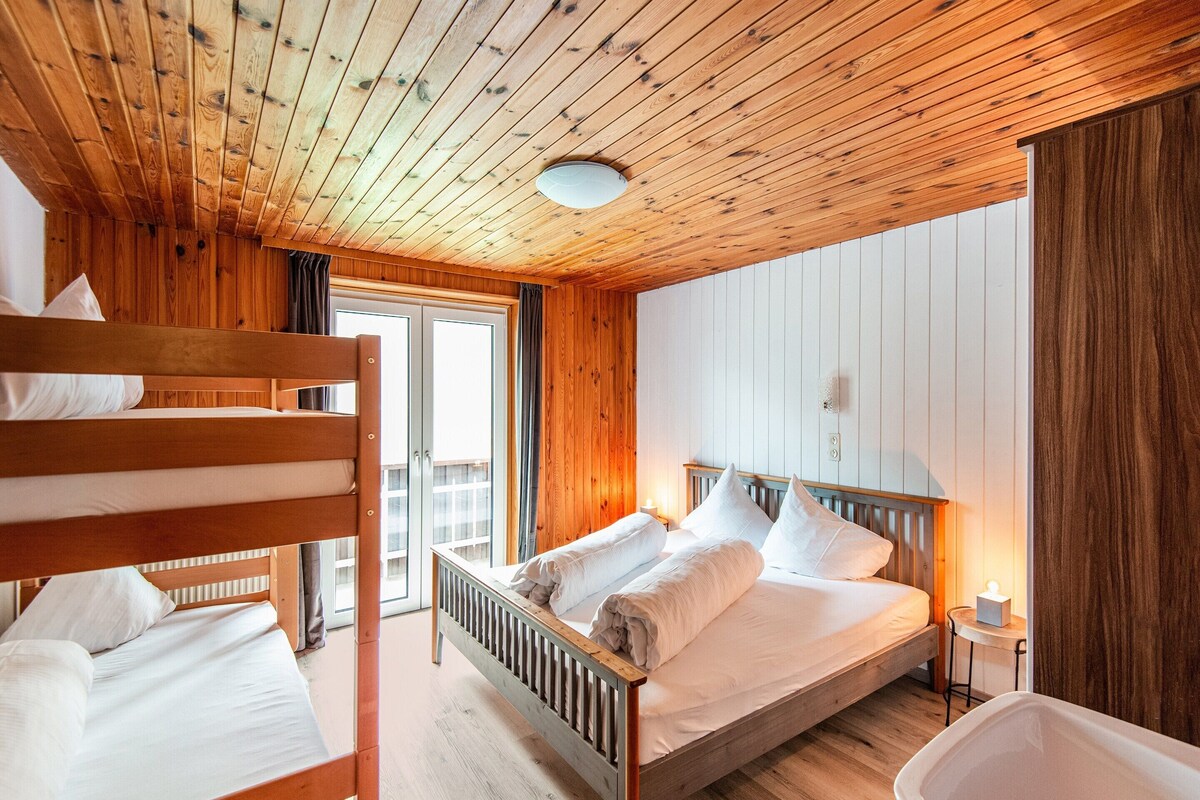A wooden cabin room with bunk beds, a single bed, a bedside table, and a window.